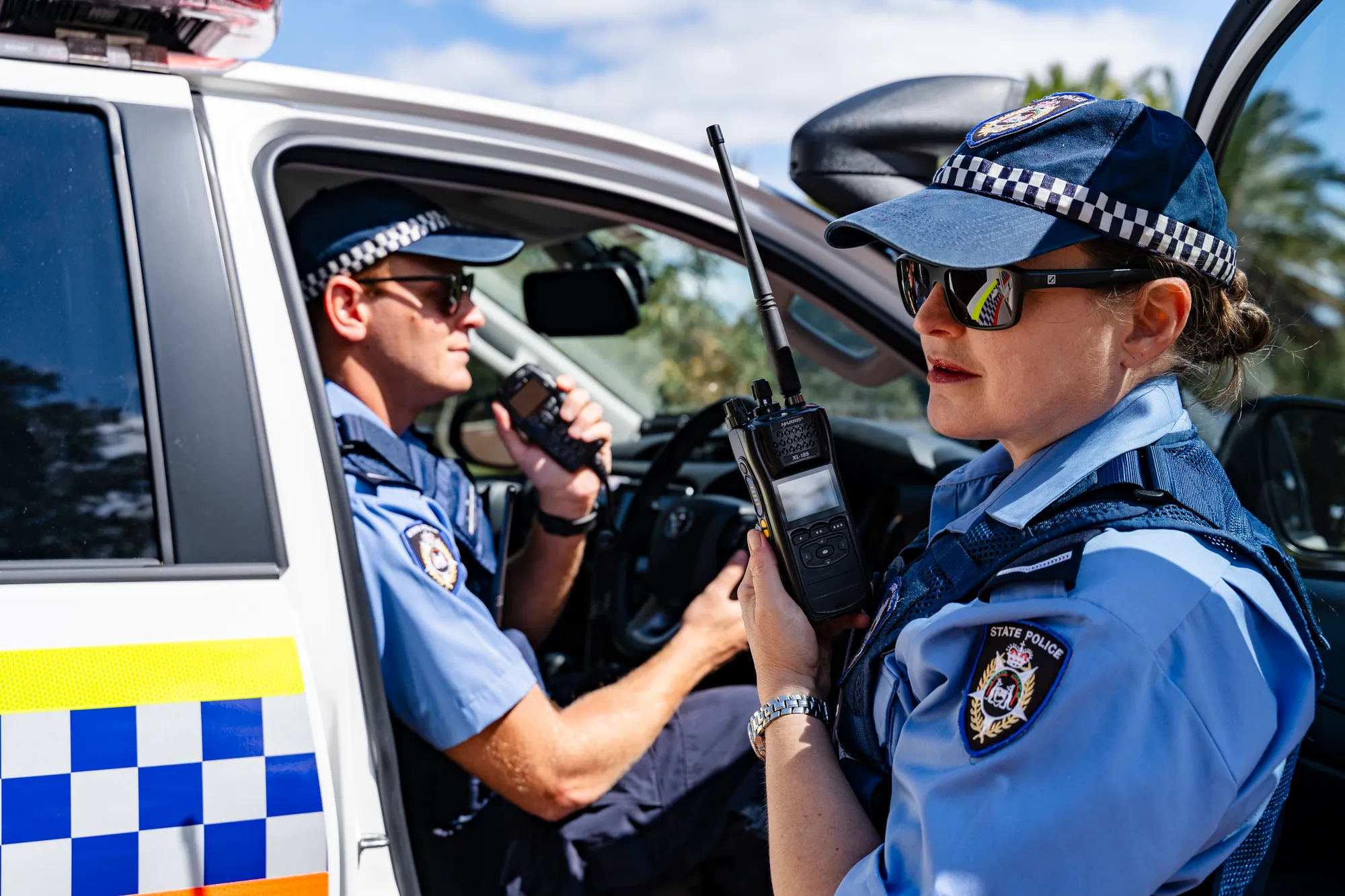 Officers in a marked police vehicle, one speaking into an L3Harris handheld radio