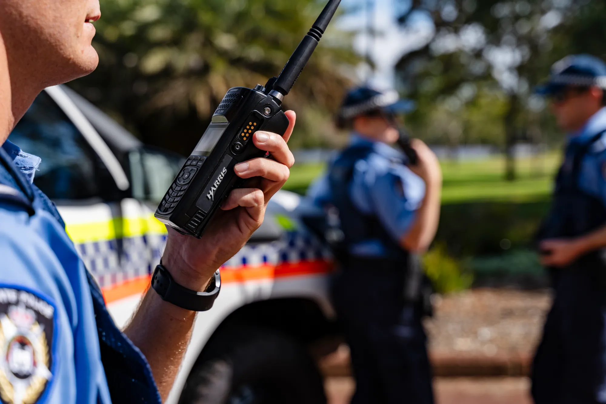 Close-up of an L3Harris handheld radio held by an officer, colleagues behind