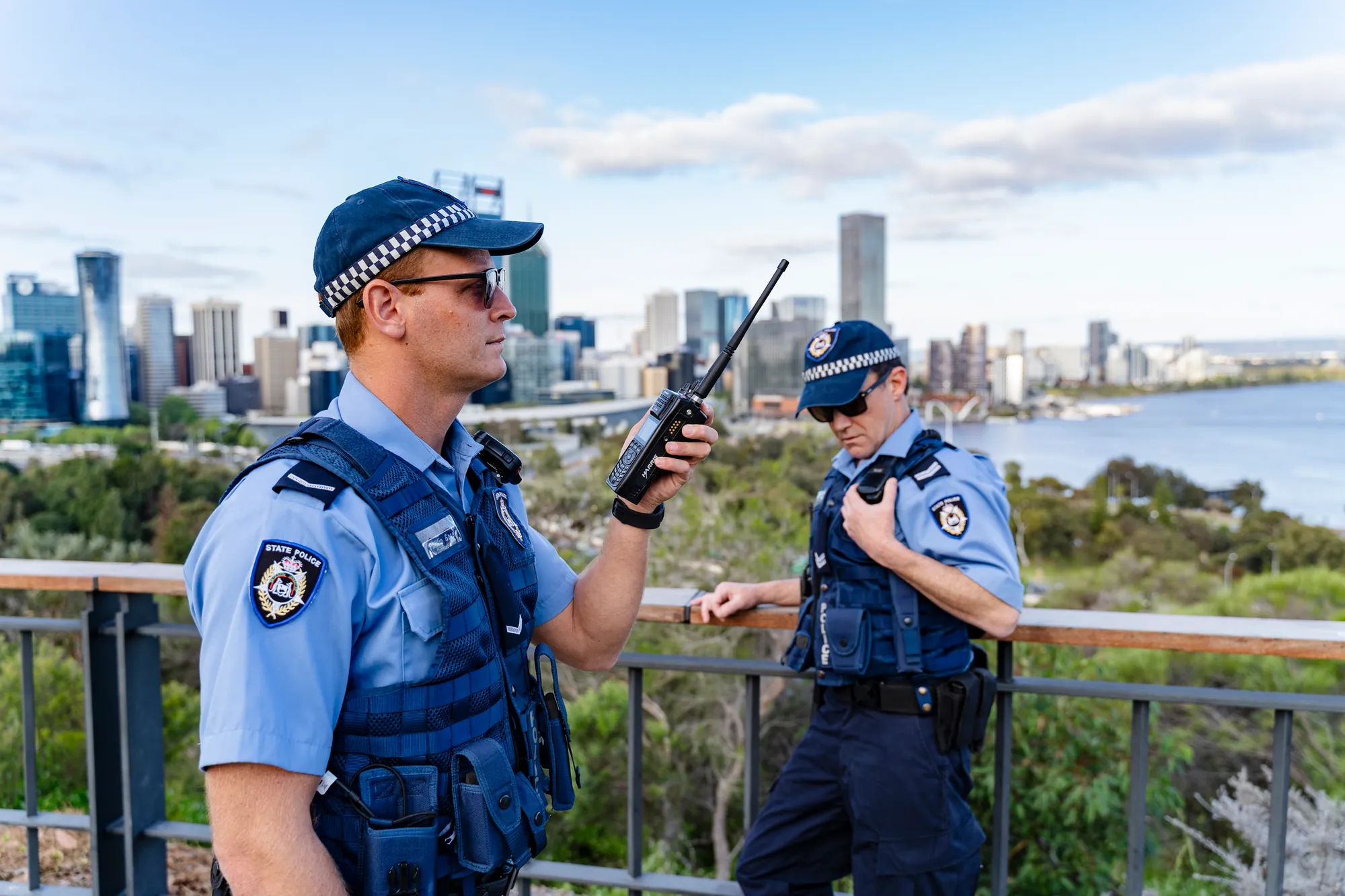 Two WA Police officers using an L3Harris handheld radio, the Perth skyline behind them