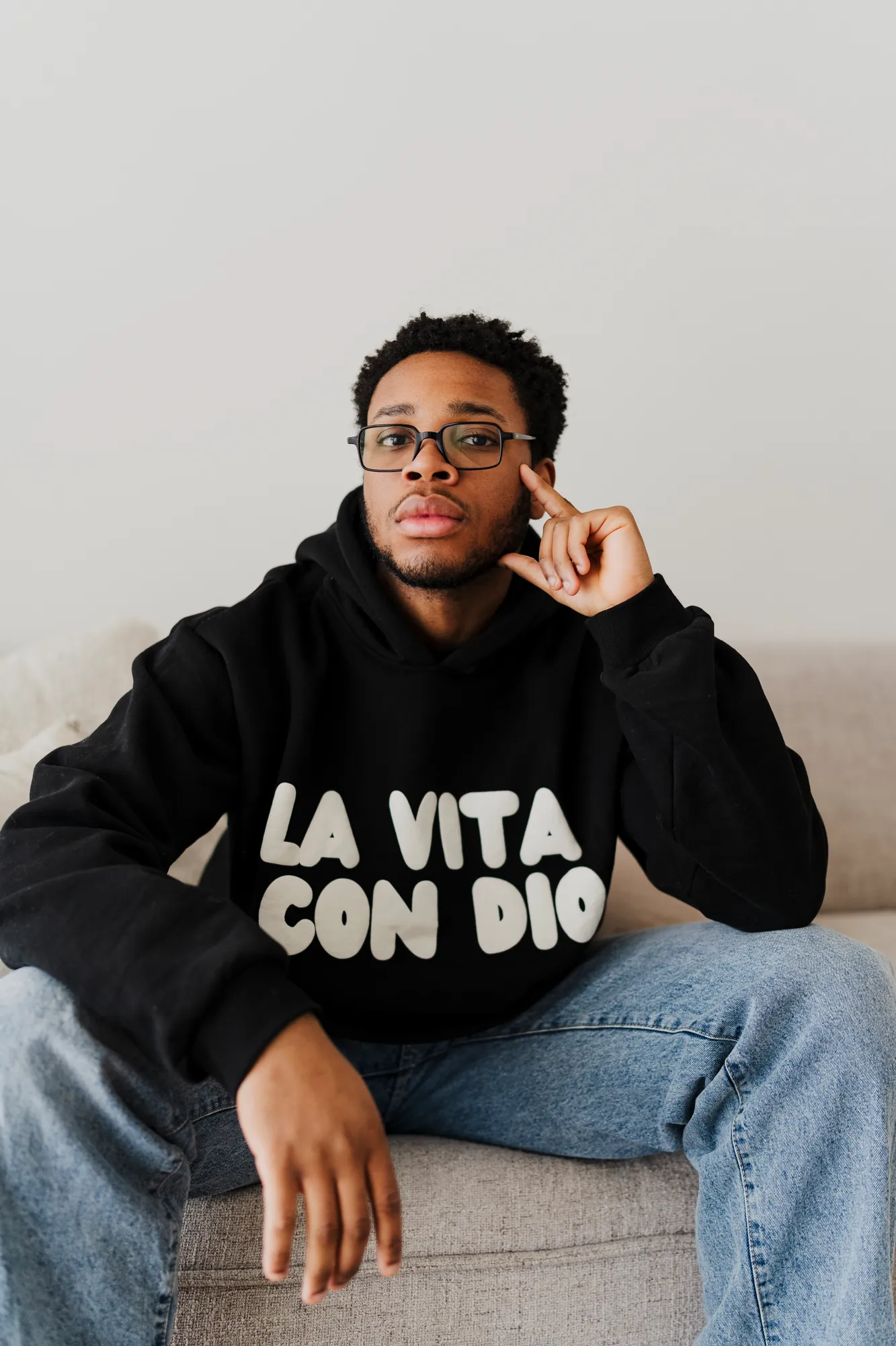 La Vita Con Dio black hoodie worn seated against a pale studio wall