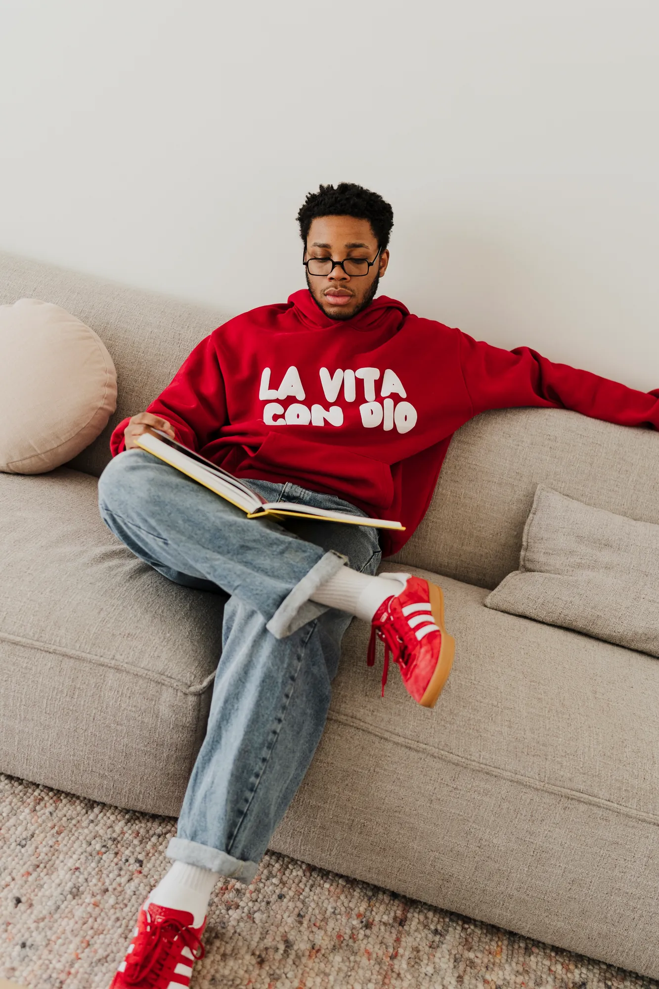 La Vita Con Dio red hoodie worn on a sofa with a book and red sneakers
