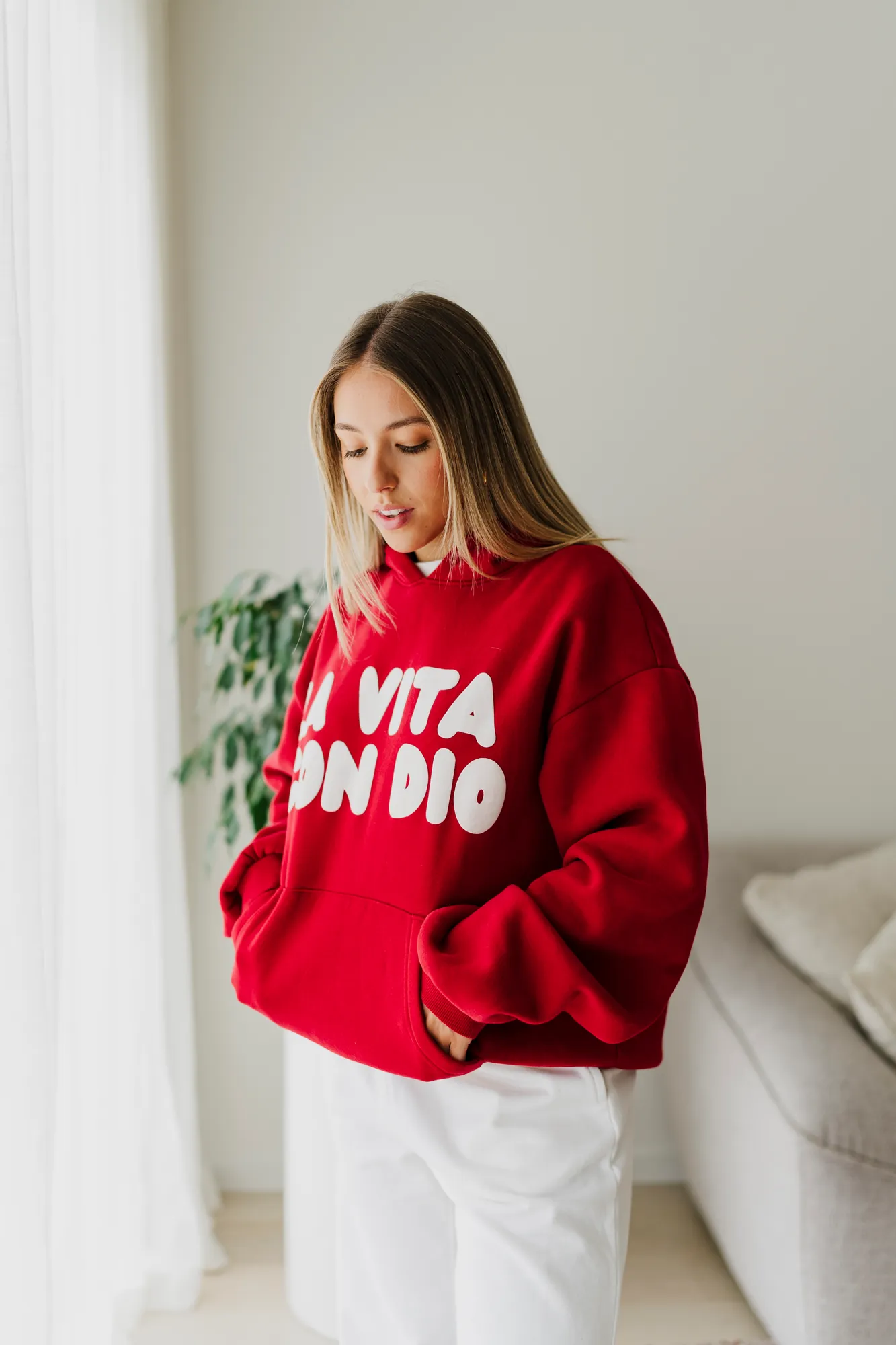 La Vita Con Dio red crew worn beside a window and a linen curtain