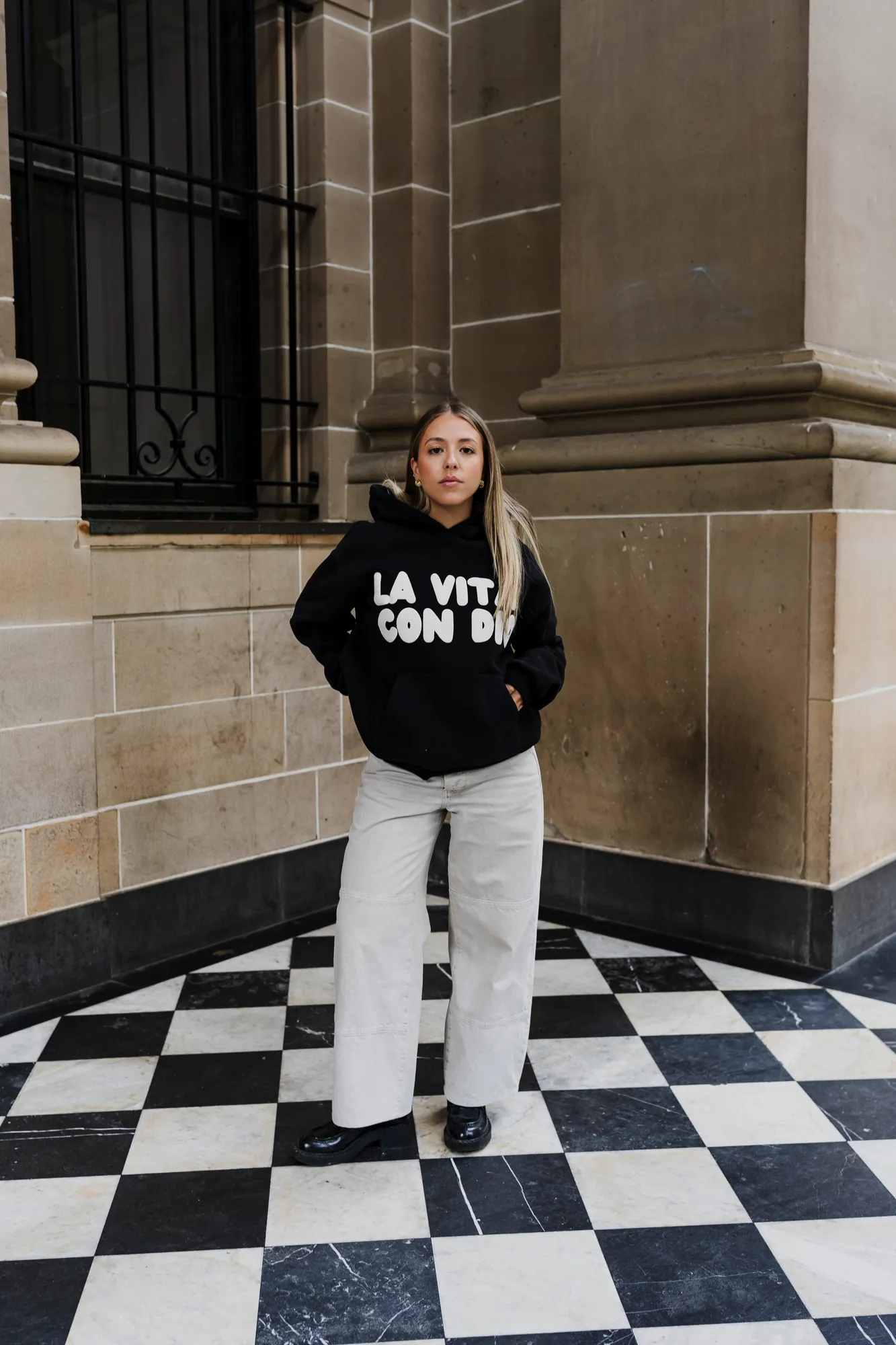 La Vita Con Dio black hoodie on a chequered tile floor outside a stone building
